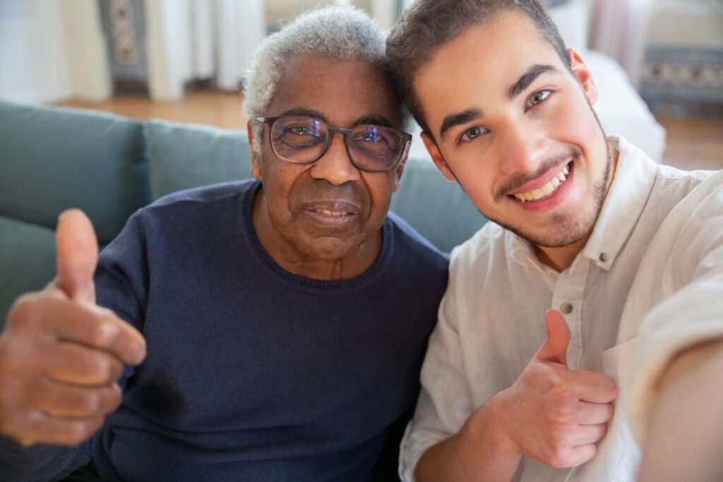 An elderly man and a young man smiling with thumbs up at home, representing friendship and care.